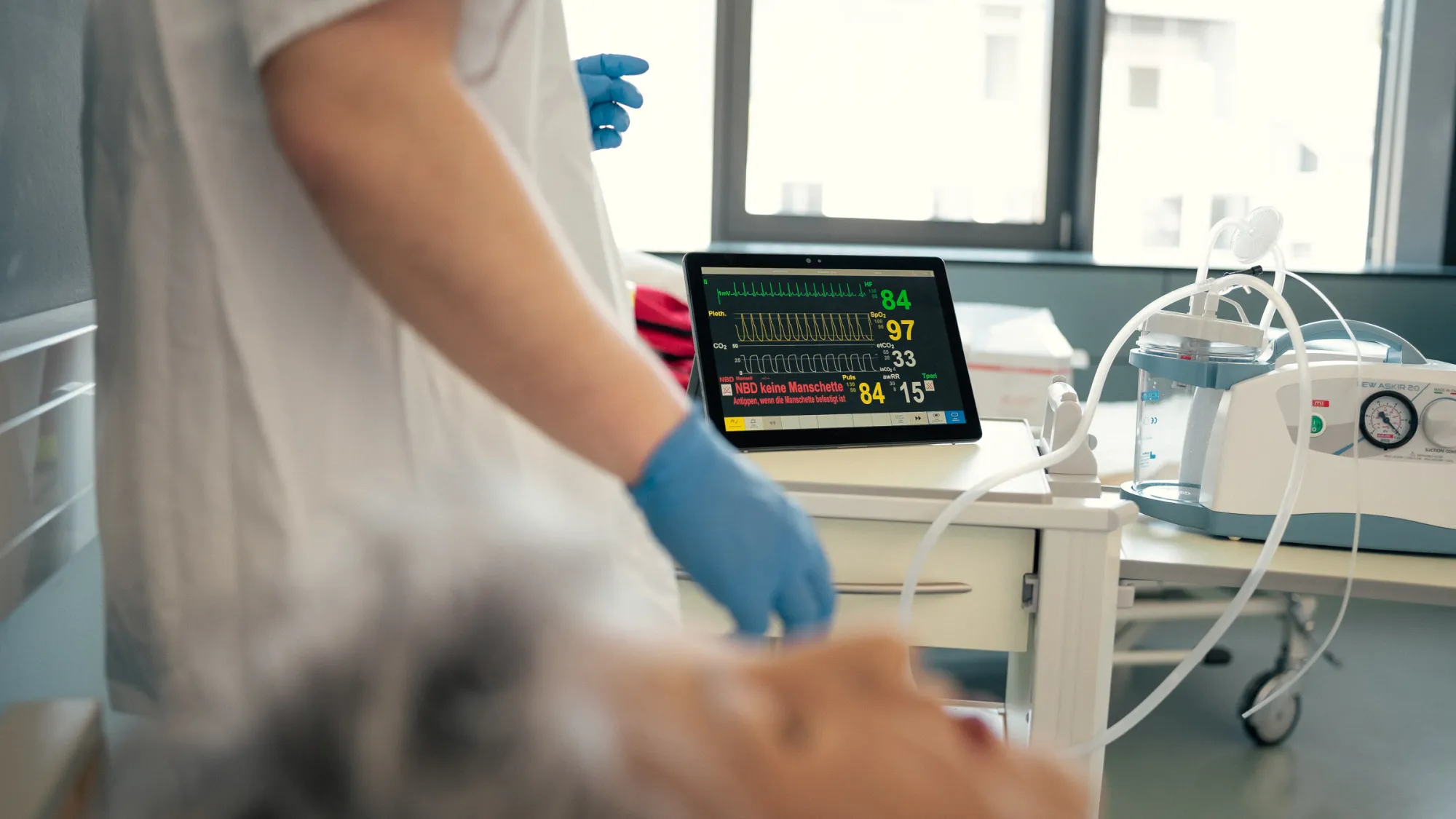 Ein medizinischer Monitor zeigt Vitalwerte an, während eine Pflegekraft mit blauen Handschuhen neben einer Übungspuppe arbeitet.