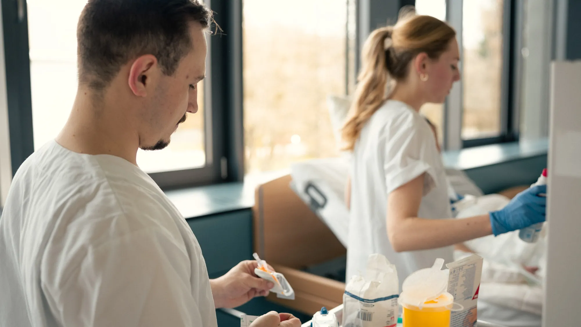 Zwei Personen in weißer Arbeitskleidung bereiten medizinische Utensilien in einem Pflegezimmer vor. Im Hintergrund steht ein Bett.