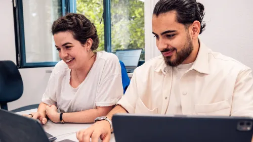 Nahaufnahme von zwei Studierenden schauen lächelnd auf ihre Laptops und Tablets. 