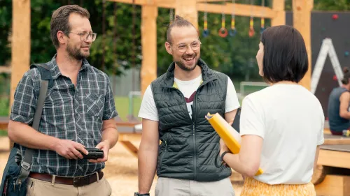Drei Personen stehen vor einem Hangelgerüst auf einer Wiese. 