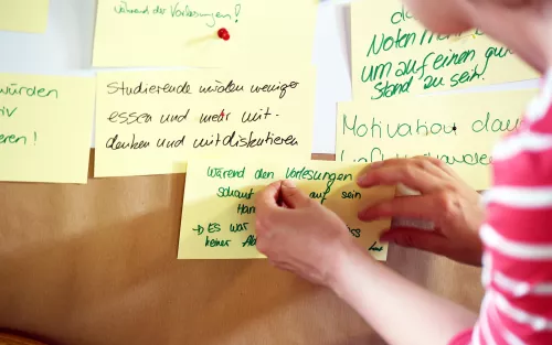 Eine Studierende pint eine beschriebene Moderationskarte an eine Stellwand der Qualitätswerkstatt