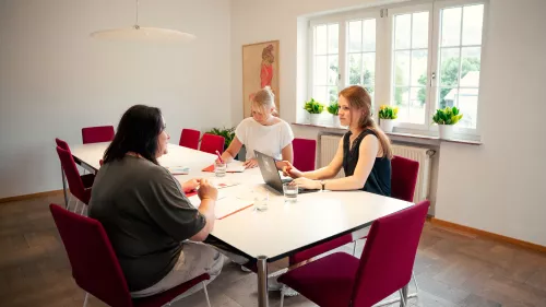 Drei Personen sitzen an einem Tisch mit Unterlagen und Laptop an einem Tisch. Eine Person schreibt, die anderen beiden sprechen miteinander.