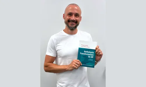 Ein Mann in einem weißen T-Shirt hält lächelnd ein Buch in die Kamera.  