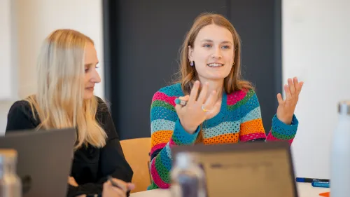 Zwei junge Frauen sitzen mit Laptops an einem Tisch. Eine Frau im bunten Pullover spricht gestikulierend.