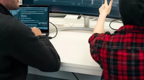 Blick über die Schultern von zwei Personen, die auf einen Computerbildschirm mit Quellcodes schauen. Halbnahe Einstellung.