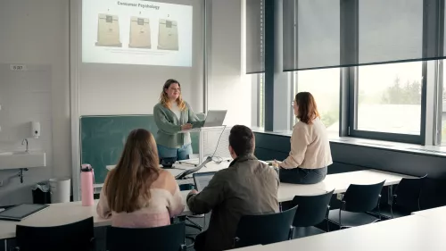 Eine Studierende im Fokus mit einer PowerPoint-Präsentation hinter ihr an der Wand, drei Studierende sitzen an und auf Tischen und schauen auf die Präsentation