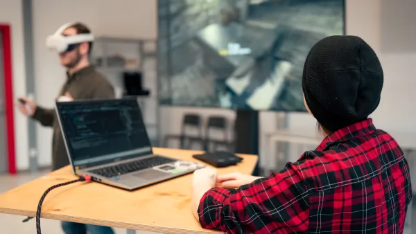 Eine Person steht im Vordergrund am Laptop. Eine weitere Person im Hintergrund trägt eine Virtual-Reality-Brille sowie einen Controller und demonstriert die Nutzung. 