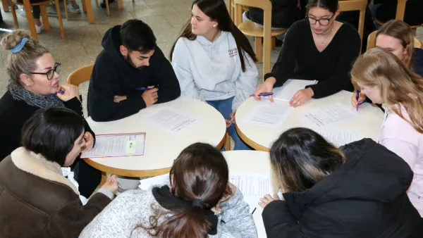Studierende sitzen in einer Runde an mehreren Tischen und bearbeiten gemeinsam Aufgabenblätter.