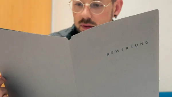 Halbnahe Aufnahme einer Person mit einer Bewerbungsmappe in der Hand an einem Schreibtisch. 