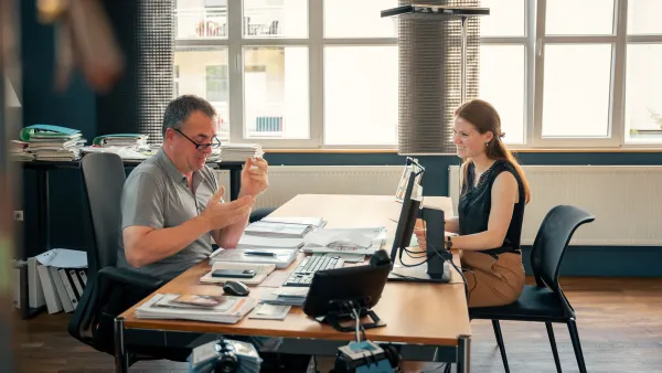 Zwei Personen sitzen an einem Tisch mit Unterlagen und Büromaterial und sprechen miteinander.