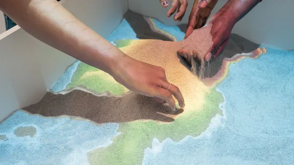 Anschnitt von zwei internationalen Studierenden, die in einem großen Kasten mit einer beleuchteten Sandkarte des International Office mit den Händen Sand verschieben.