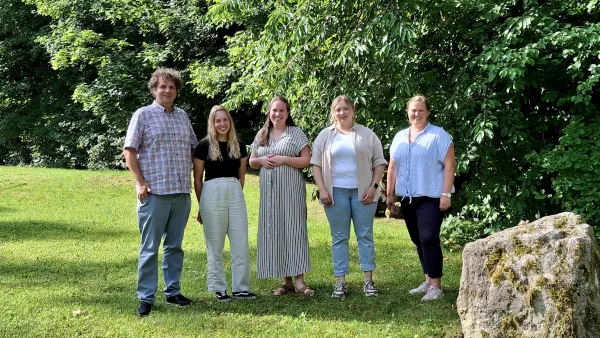 Gruppenfoto von fünf Personen auf einer grünen Wiese, rechts im Bild liegt ein Stein und im Hintergrund ist ein Baum. 