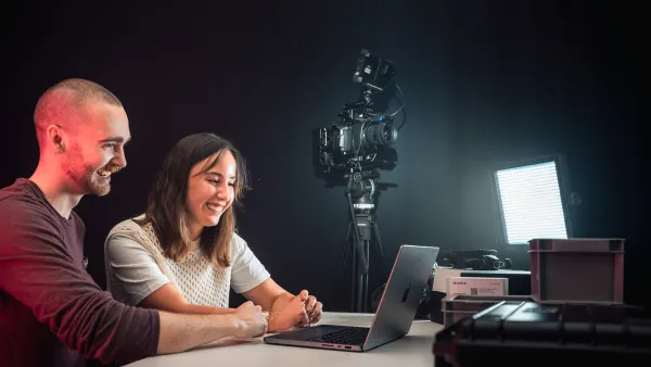 Zwei Studierende arbeiten an einem Laptop. Im Hintergrund stehen eine Kamera und eine Lampe.