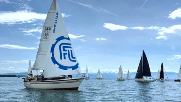 Das Segelboot des RWU-Segelclubs auf dem Bodensee, im Hintergrund sind viele weitere Boote und die Berge zu sehen.