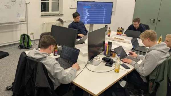 Sechs Mitglieder des Teams „Weingarten Wiresharks“ sitzen konzentriert an einem großen Tisch und arbeiten an Laptops und Monitoren. Ein Whiteboard mit Notizen, Getränke und Pizza-Kartons auf dem Tisch zeigen die Arbeitsatmosphäre während des laufenden Events.