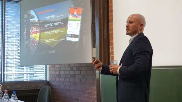 Referent Wieland Schwarze im dunkelblauen Sakko steht während seines Gastvortrags an der RWU vor einer projizierten Präsentationsfolie, die die Benutzeroberfläche der Mainz 05 App auf einem Smartphone sowie strategische Kernpunkte zur App-Optimierung zeigt.