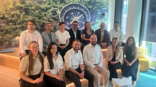 Gruppenbild in einem hellen Büro: Eine Gruppe von Studierenden und Dozenten sitzt lächelnd in zwei Reihen auf einer holzfarbenen Treppenstufe. Im Hintergrund befindet sich eine große Wandtapete, die eine Waldlandschaft mit einem zentralen Kompass-Motiv zeigt. Rechts unten auf dem Boden liegt ein weißes Kissen mit dem goldgas-Logo.