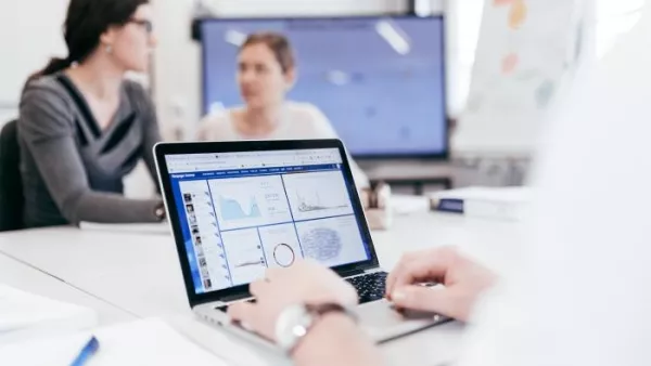 Eine Person am Laptop mit Datenvisualisierungen auf dem Bildschirm, im Hintergrund sind zwei Frauen im Gespräch.