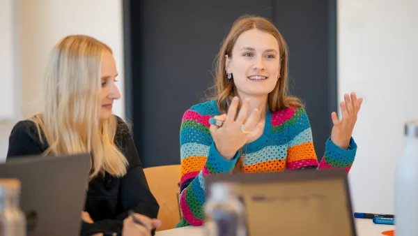 Zwei junge Frauen sitzen mit Laptops an einem Tisch. Eine Frau im bunten Pullover spricht gestikulierend.