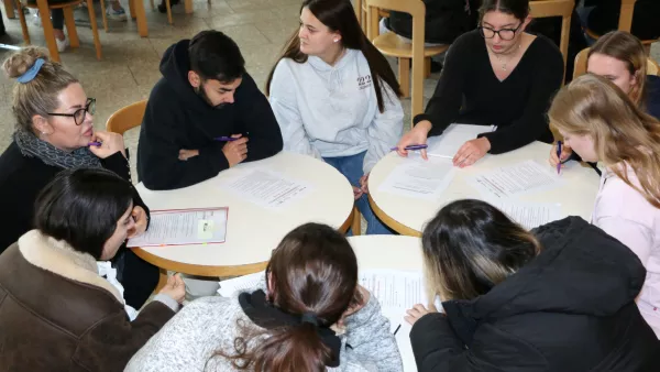 Studierende sitzen in einer Runde an mehreren Tischen und bearbeiten gemeinsam Aufgabenblätter.