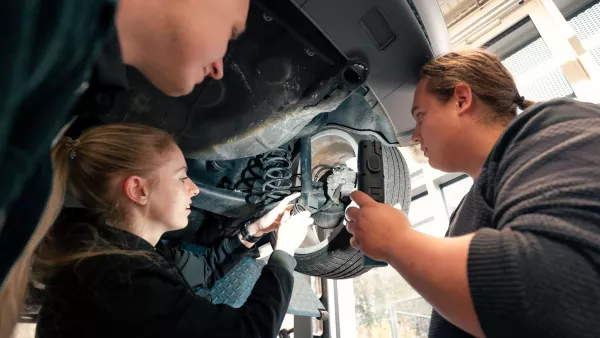 Blick von unten auf Studierende, die ein Auto von unten betrachten. Einer hält eine Taschenlampe, eine Studentin hält ein Bauteil in der Hand. Das Auto ist auf einer Hebebühne.