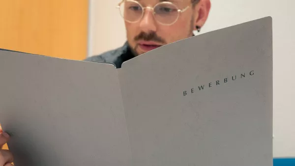 Halbnahe Aufnahme einer Person mit einer Bewerbungsmappe in der Hand an einem Schreibtisch.