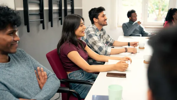 Seitenansicht, halbnah: Mehrere Studierende sitzen an Tischen in einem Seminarraum des International Office. Sie lächeln. Eine Studierende ist im Fokus der Kamera.