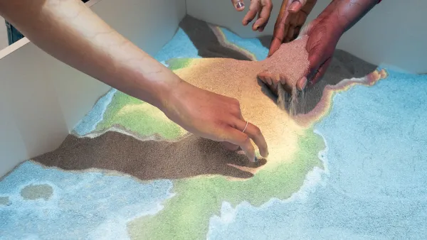 Anschnitt von zwei internationalen Studierenden, die in einem großen Kasten mit einer beleuchteten Sandkarte des International Office mit den Händen Sand verschieben.