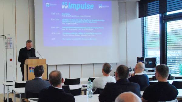 Redner an Pult vor einem Hörsaal, auf der Leinwand die Überschrift "IDW Impulse".