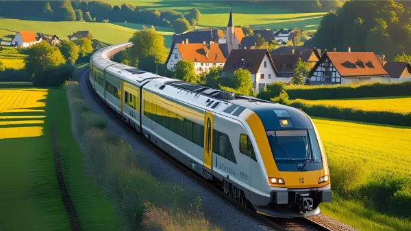Ein Regionalzug fährt durch eine grüne Landschaft. Im Hintergrund sind Häuser und Bäume zu sehen