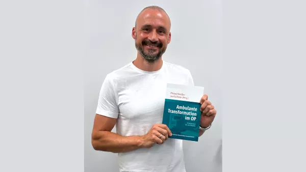 Ein Mann in einem weißen T-Shirt hält lächelnd ein Buch in die Kamera.