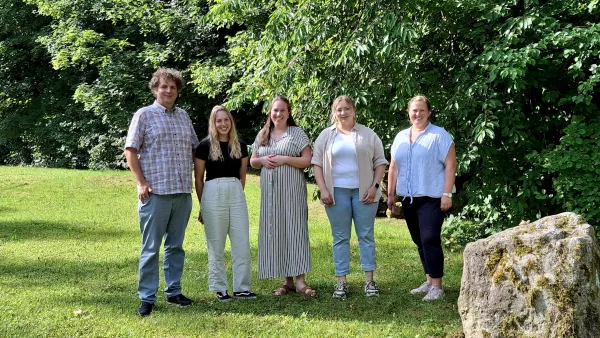 Gruppenfoto von fünf Personen auf einer grünen Wiese, rechts im Bild liegt ein Stein und im Hintergrund ist ein Baum.