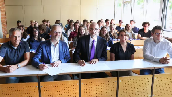 Fünf Personen sitzen in der ersten Reihe eines Hörsaals und unterzeichnen den Kooperationsvertrages mit dem Welfen-Gymnasium. Im Hintergrund sind Schülerinnen und Schüler.