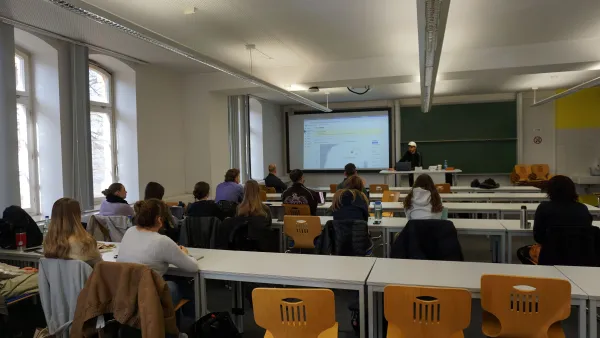 Ein Referent steht vor einer großen Leinwand in einem Hörsaal und hält einen Praxisvortrag. Auf der Leinwand ist das Dashboard einer SAP-Software für Marketing Automation mit verschiedenen Datenanalysen und Menüpunkten zu sehen. Studierende sitzen an Tischen im Vordergrund und verfolgen die Präsentation.