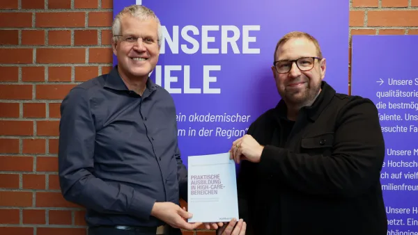 Prof. Dr. Jörg Wendorff und Georg Roth mit dem Buch "Praktische Ausbildung in High-Care-Bereichen" vor den Leitbildern der RWU.