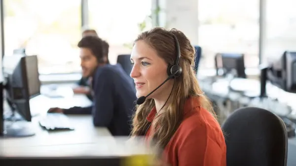 Eine junge Frau mit Headset sitzt konzentriert an einem PC-Arbeitsplatz in einem hellen Großraumbüro, während im Hintergrund ein weiterer Kollege am Schreibtisch telefoniert und am Monitor arbeitet.