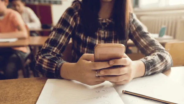 Eine Nahaufnahme, die auf die Hände und das Smartphone einer jungen Person in einem Klassenzimmer fokussiert ist. Die Person, die ein kariertes Flanellhemd trägt und deren Gesicht nicht vollständig sichtbar ist, sitzt an einem Holztisch und blickt mit gesenktem Kopf konzentriert auf den Bildschirm des Telefons, das sie in beiden Händen hält. Ihre Hände befinden sich direkt über einem offenen, blanken Notizbuch. Im unscharfen Hintergrund sitzt ein weiterer Schüler an einem Tisch, und warmes Sonnenlicht fällt