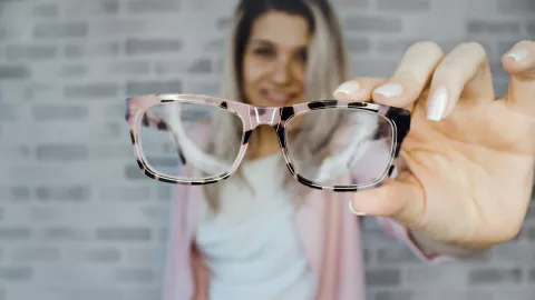 Eine junge Frau hält lächelnd eine Brille in die Kamera
