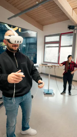 Eine Person trägt eine Virtual-Reality-Brille sowie einen Controller und demonstriert die Nutzung. Eine Person schaut zu. Im Hintergrund ist ein großer Bildschirm zu sehen.